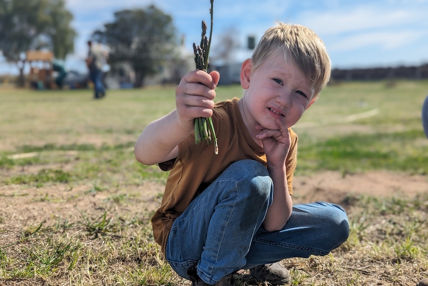 A young blonde-boy, Lane, holds a bunch of freshly picked green asparagus as he squints into the sun.