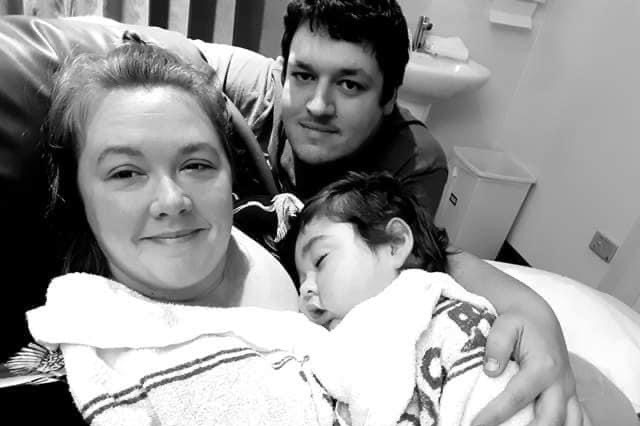 A black and white photo of a woman lying on a hospital bed holding a young boy on her chest with a man crouched behind them.