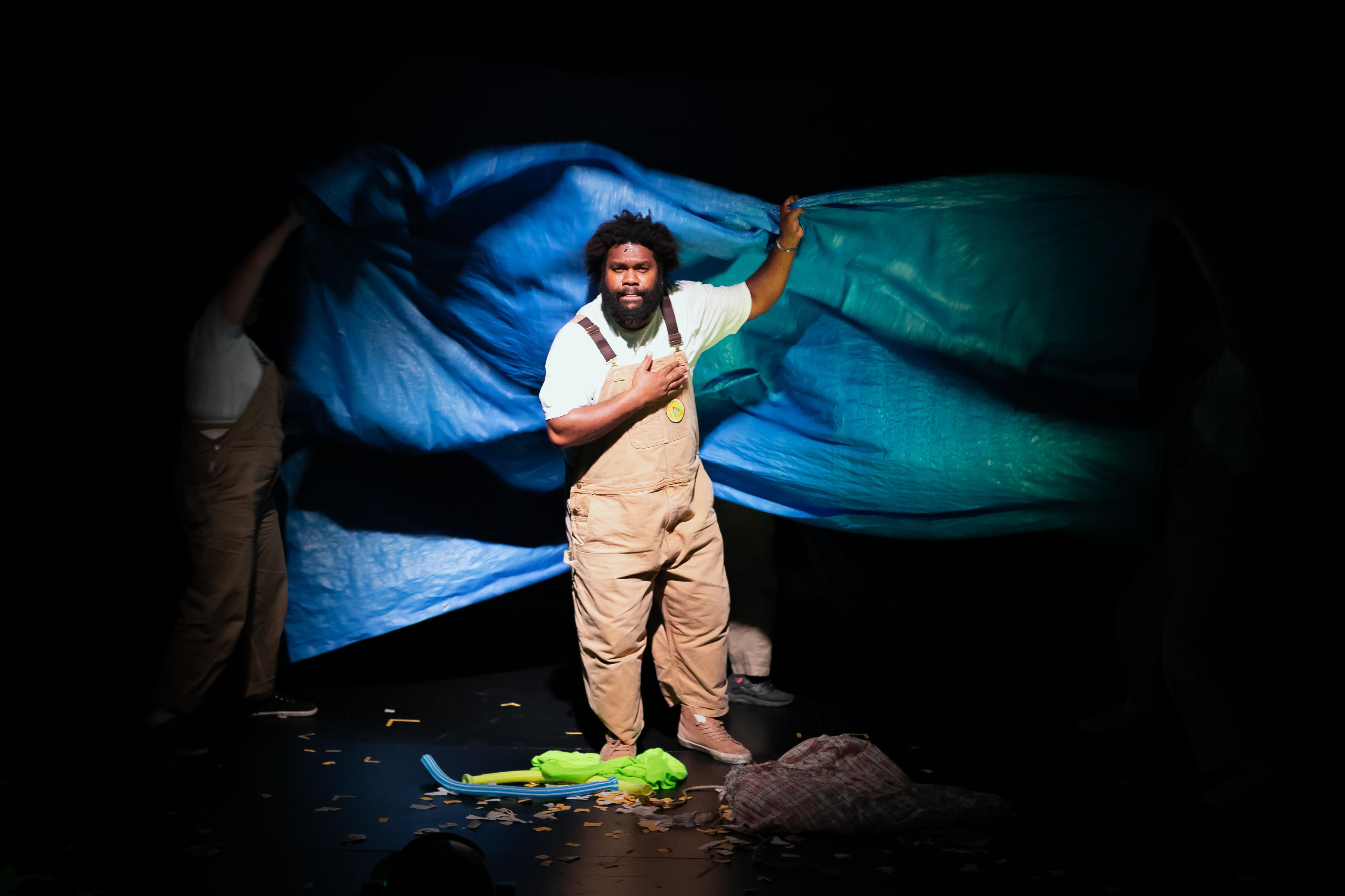 Indigenous man wearing caramel overalls stands on a darkened stage with his hand to his heart and flanked by a blue tarp.