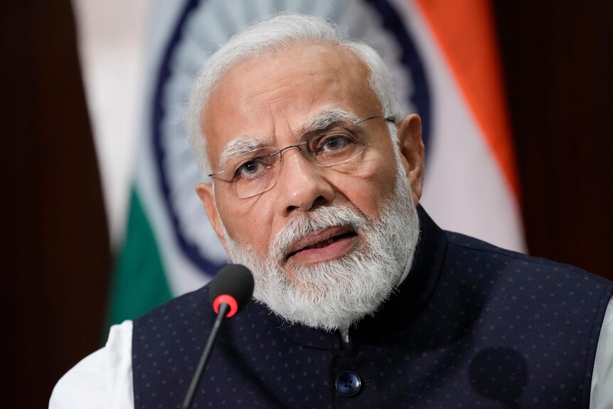 Narendra Modi wears spectacles as he leans into a microphone while he speaks while in front of an Indian flag.