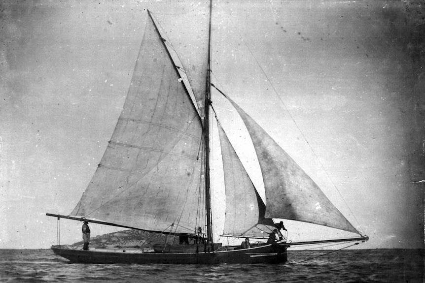 A black and white image of a fishing boat in full sail  with a small island in the background