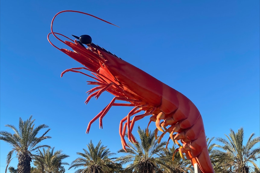 A pink prawn statue on a stick in front of a blue sky., top of palm trees in the background.