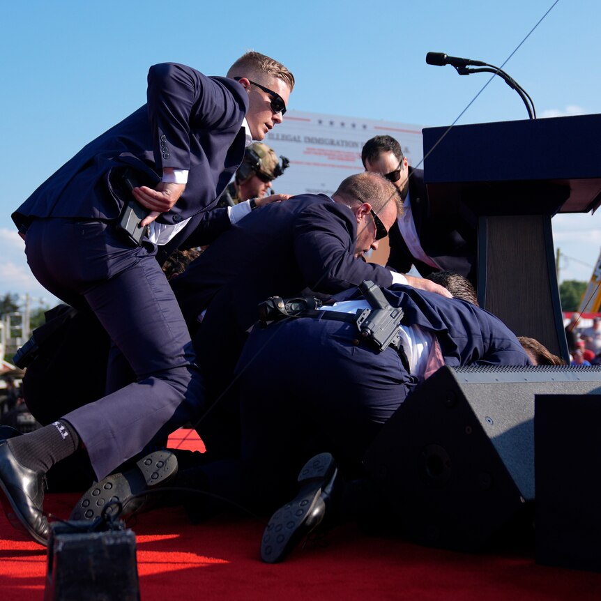 A group of officers in suits lie in a pile as seen from behind the stage. 