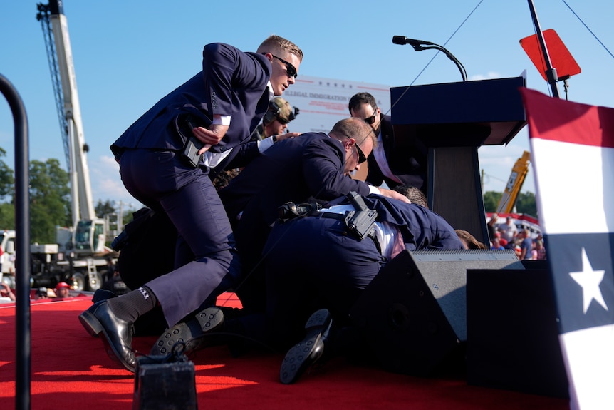 A group of officers in suits lie in a pile as seen from behind the stage. 