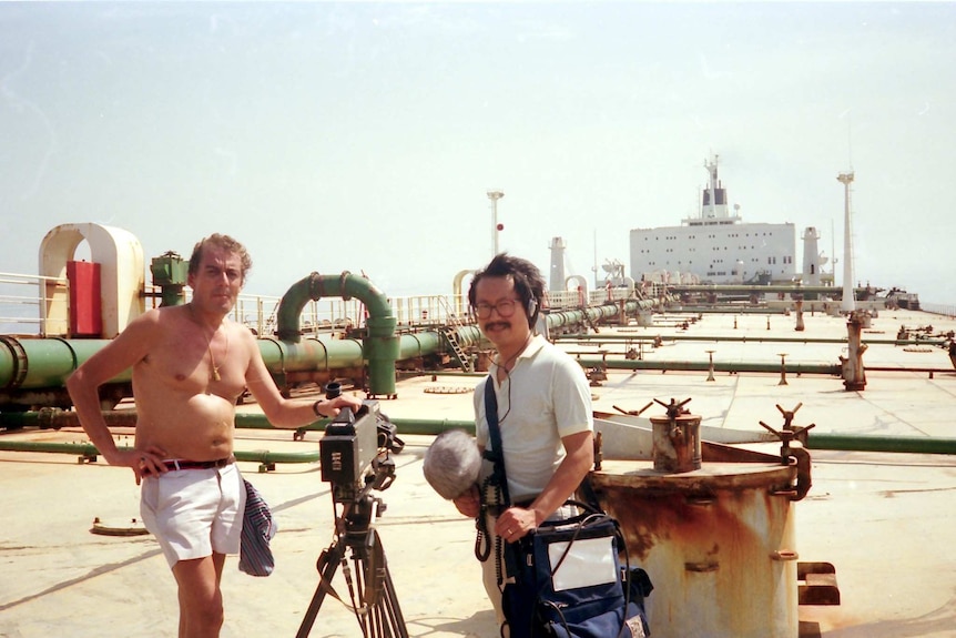 Derek Williams stands with his camera on a tanker beside a soundman holding audio equipment