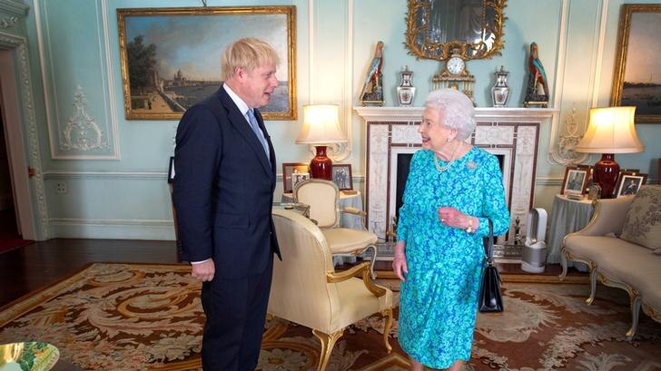 Boris Johnson stands to the left of a living room, with the Queen on the right. He wears a dark suit, she is in a blue dress.