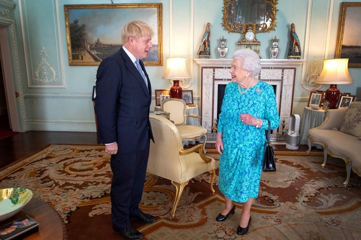 Boris Johnson stands to the left of a living room, with the Queen on the right. He wears a dark suit, she is in a blue dress.