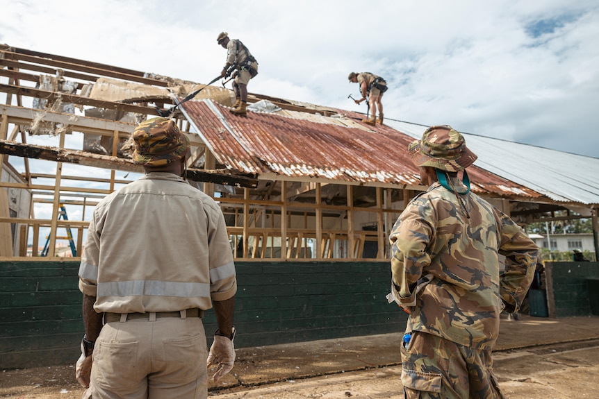 Two men in military uniforms watch as two other men work on the roof of a half-built classroom. 