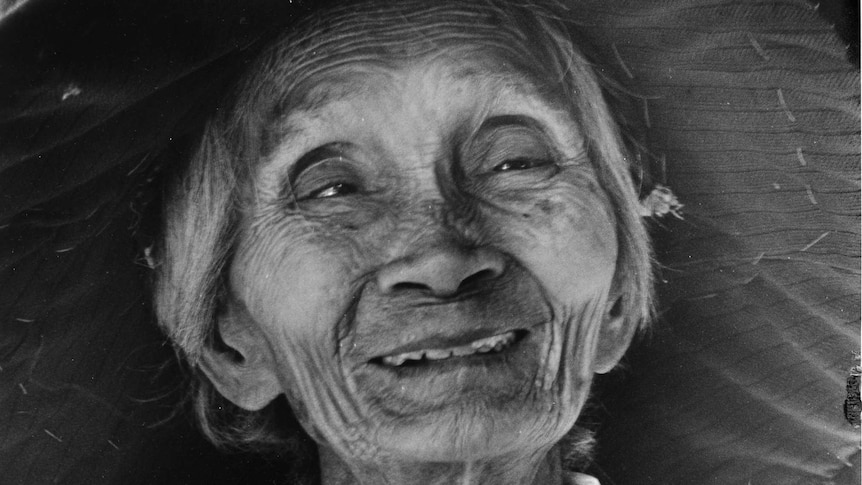 Elderly Chinese lady wearing a wide-brimmed hat crouching in her garden holding a machete looking at the camera