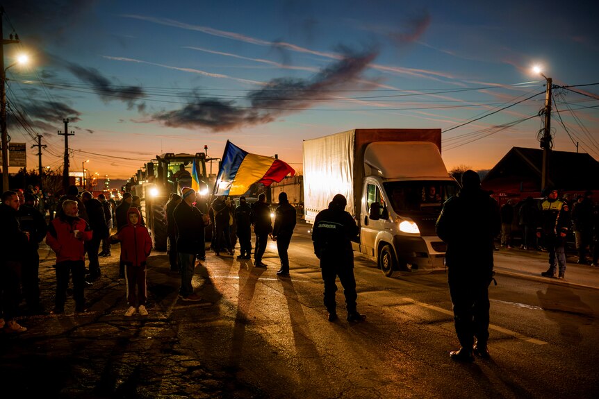 A crowd stands around trucks and tractors on the road as the sun sets.