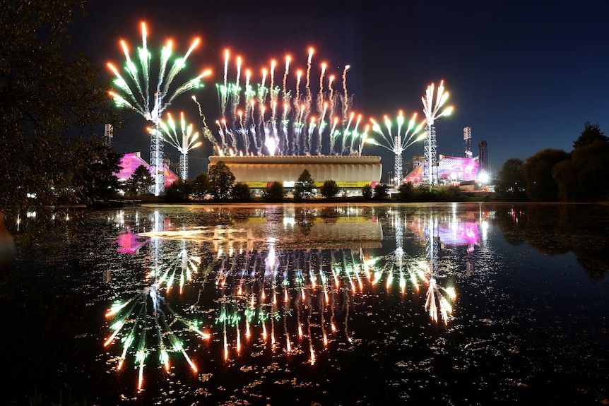 Fireworks explode above a sports stadium and reflect in a pond in front of it