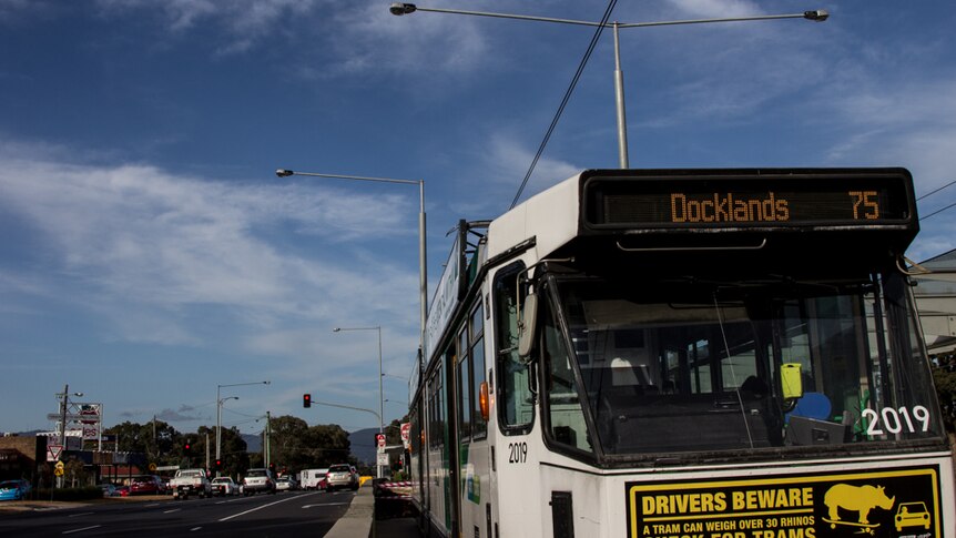 A tram at a tram stop next to a shopping centre, hills in background.