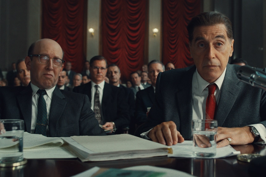 A group of white men in the American senate, two men sit at the front at a table, one (Al Pacino) sits in front of a microphone