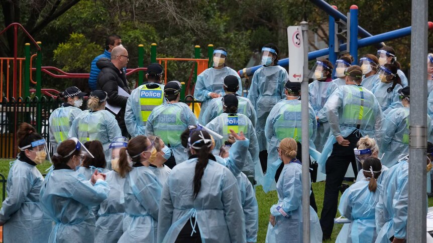 A large group of people waring blue gowns and face masks stands in front of a playground.