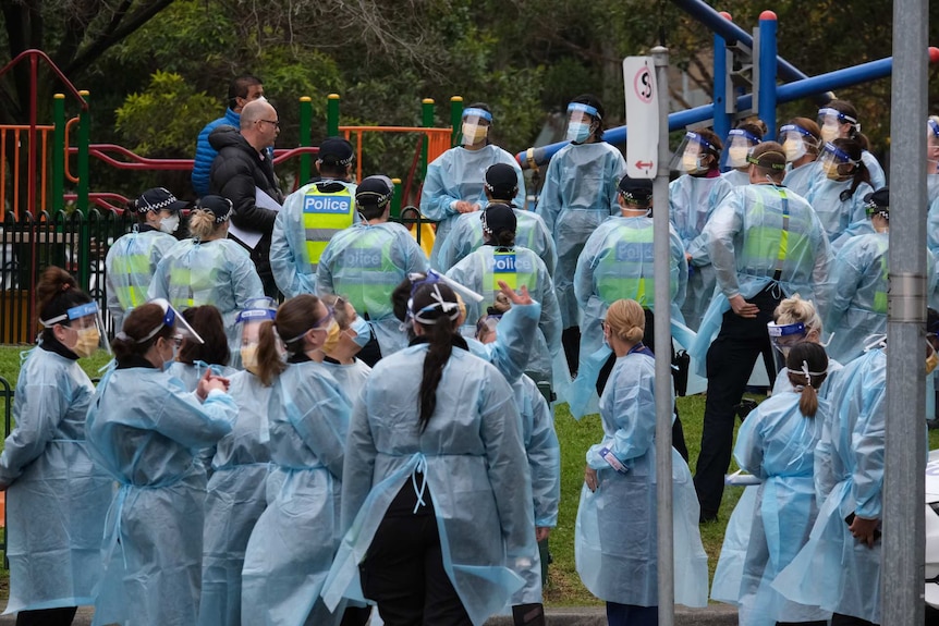 A large group of people waring blue gowns and face masks stands in front of a playground.