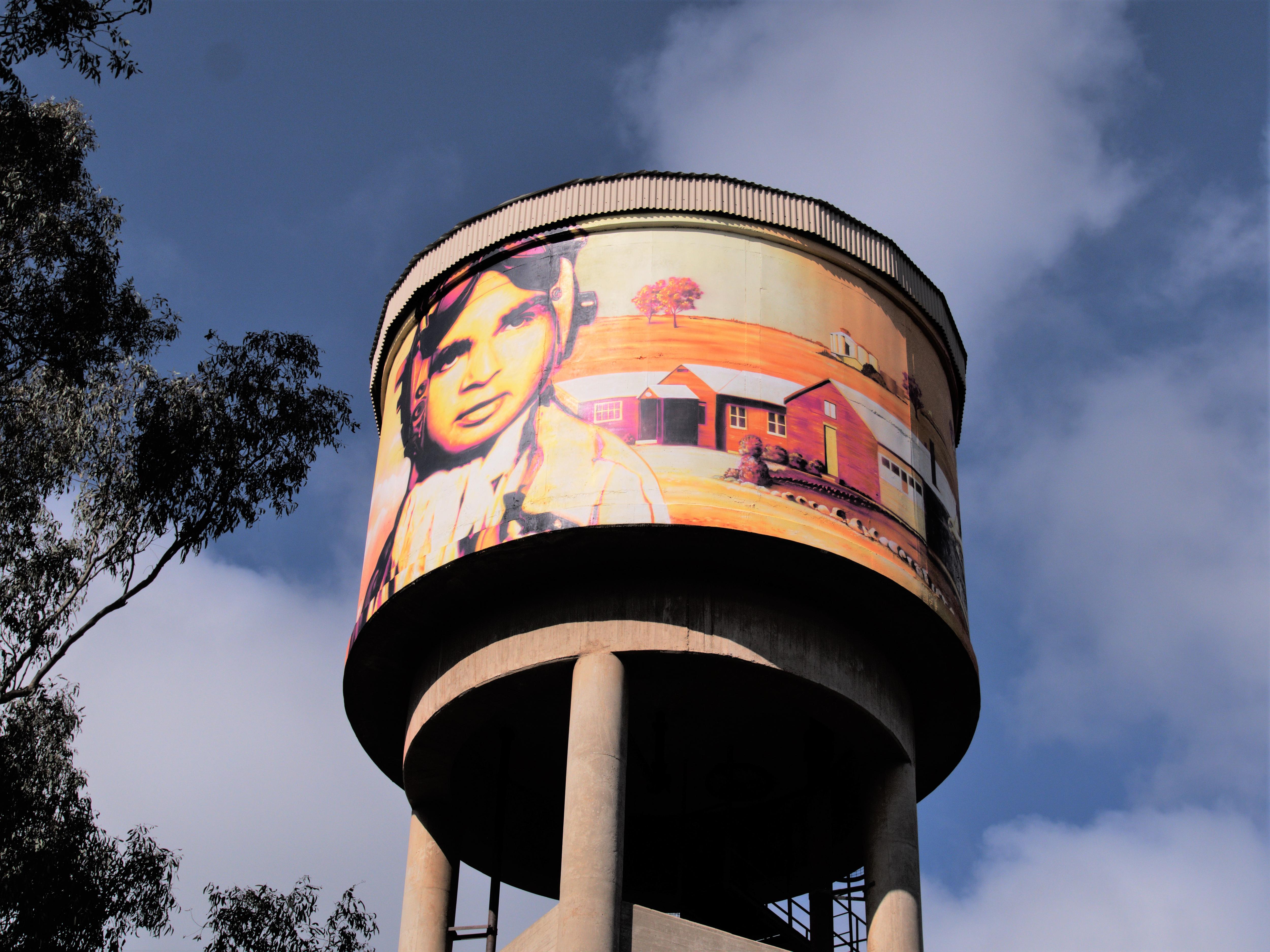 a water tower mural in bright colours with trees seen beside it.
