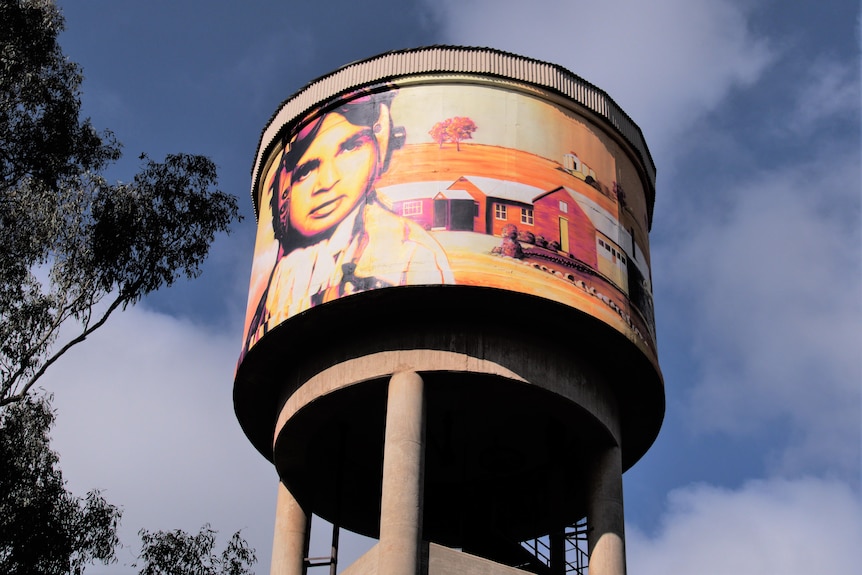 a water tower mural in bright colours with trees seen beside it.