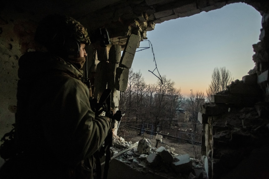 A Ukrainian serviceman looks outside a hole in a building and onto a forest.