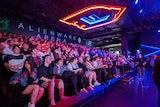 A large group of people sit on raked seating, watching an eSports event