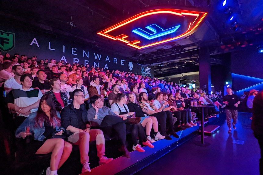 A large group of people sit on raked seating, watching an eSports event