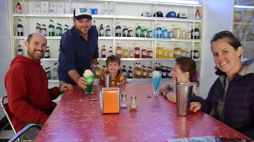 A man wearing a cap serving drinks to a smiling family in front of a shelf of bottles with different coloured liquids