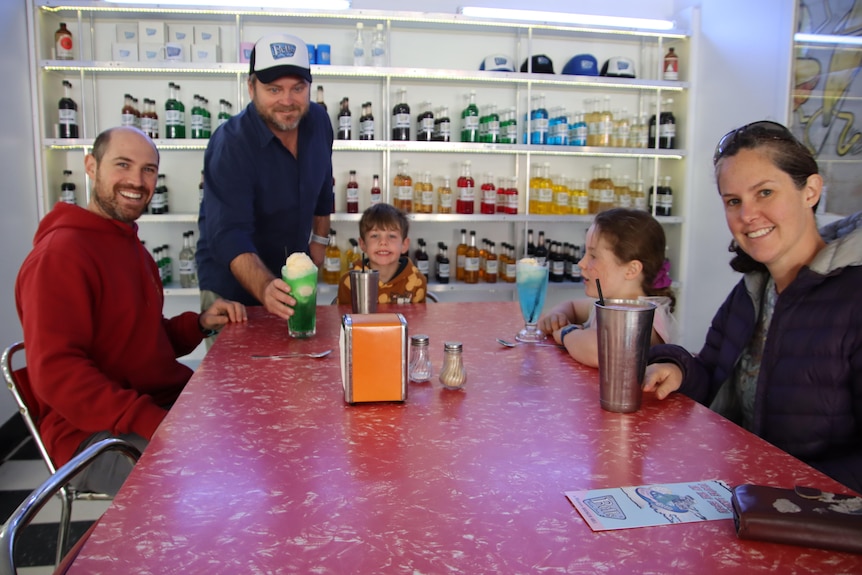 A man wearing a cap serving drinks to a smiling family in front of a shelf of bottles with different coloured liquids