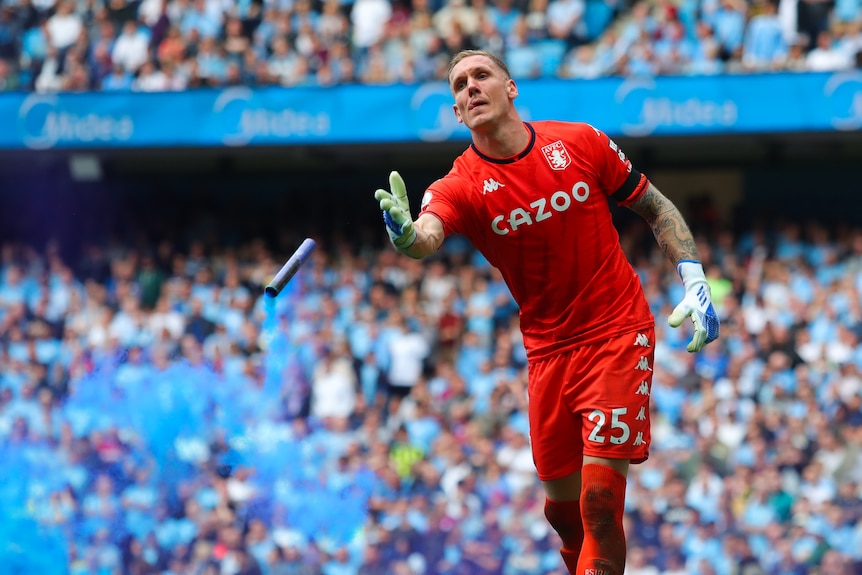 A Premier League goalkeeper dressed in red throws a canister with blue smoke away from him off the pitch during a game.