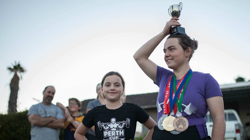 Maddy Keast stands outside her home with a trophy held above her head and medals around her neck with her sister beside her.