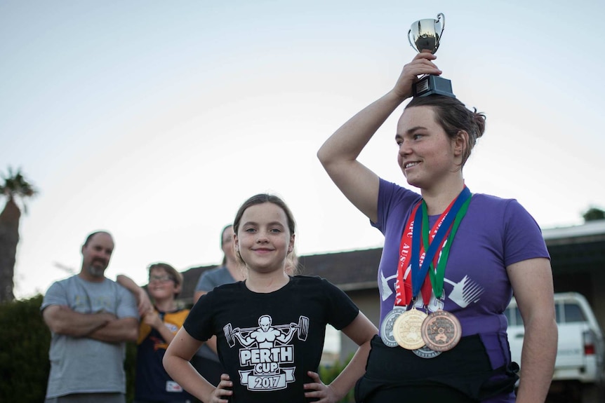 Maddy Keast stands outside her home with a trophy held above her head and medals around her neck with her sister beside her.