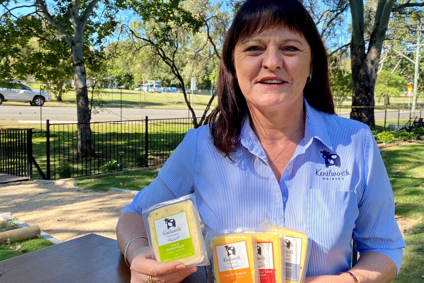 A seated woman in a blue Kenilworth Dairies shirt holds up packets of cheese