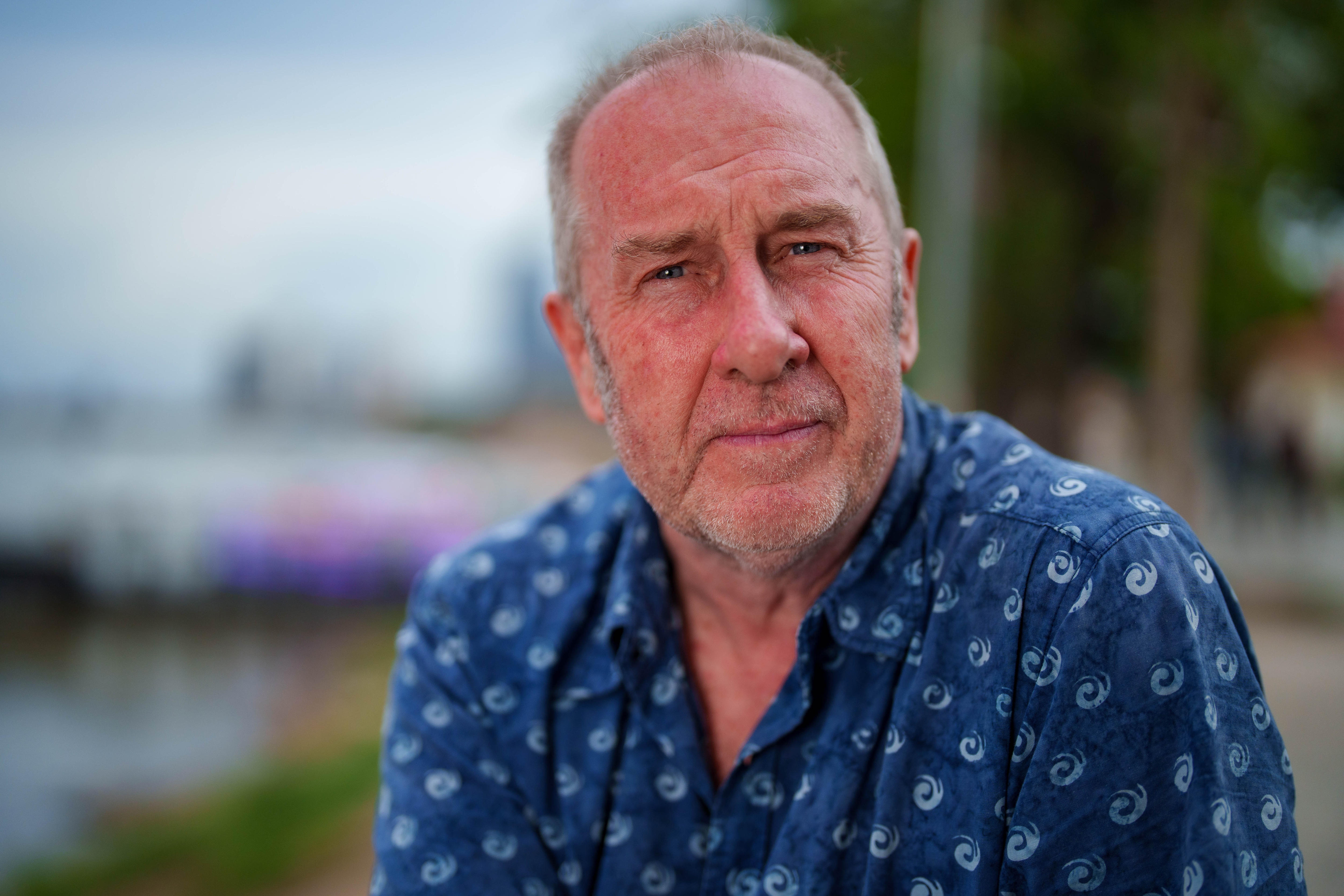 A middle-aged man in a short-sleeve blue collared shirt looks into camera with a serious expression.