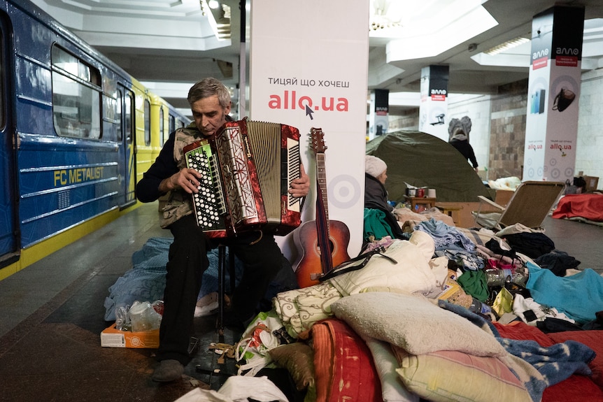 A man plays the accordion in a Kharkiv metro station, surrounded by sleeping bags and pillows