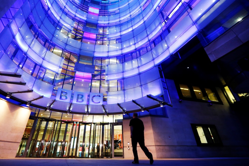 A silhouetted man walks into a brightly lit, concave modern building. The BBC logo is above the front door.