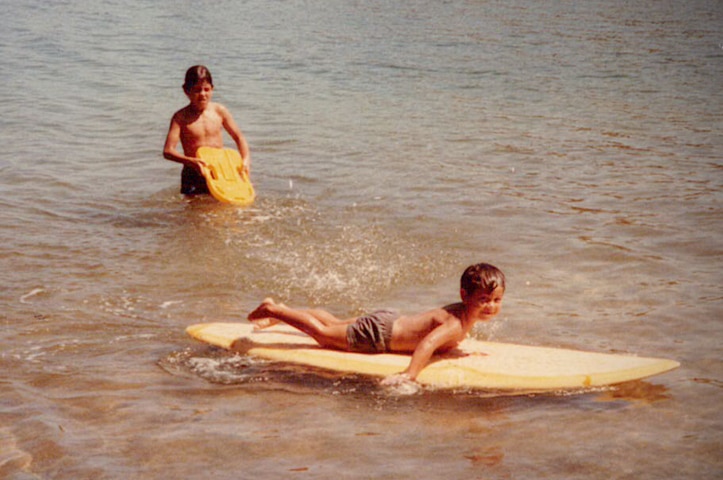 Three young boys in the water with one of them on a large surfboard.