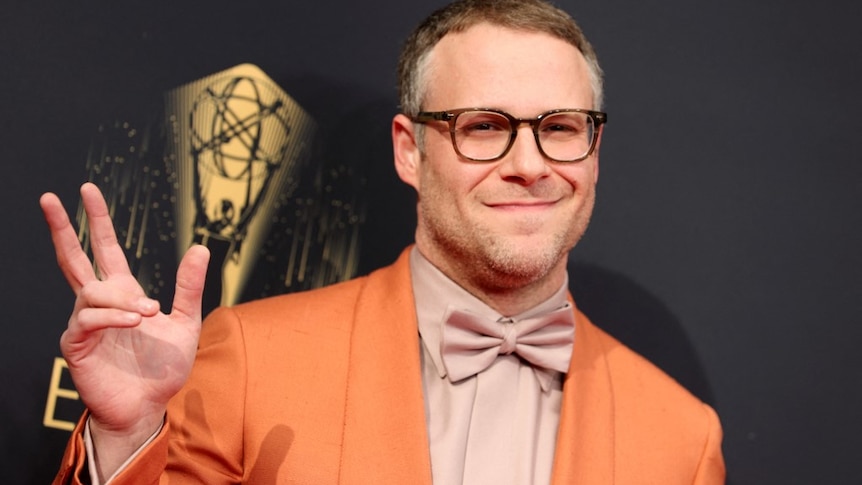 Seth Rogen gives the peace sign to the camera wearing an orange jacket and glasses