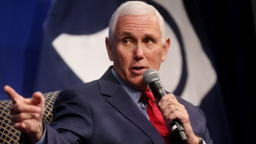 Mike Pence, wearing a blue suit and red tie, points his finger while holding a microphone up to his mouth