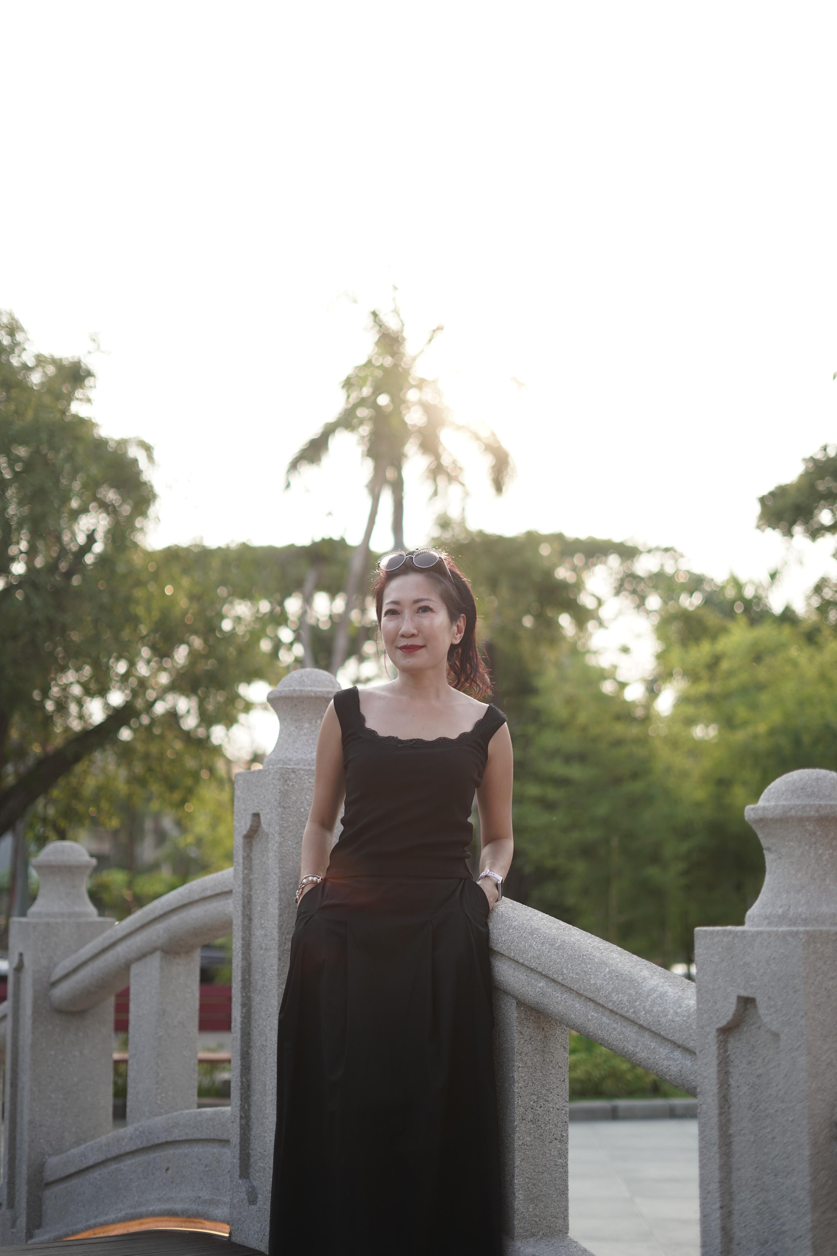 Una mujer con un vestido negro está parada en un puente con las manos en los bolsillos.