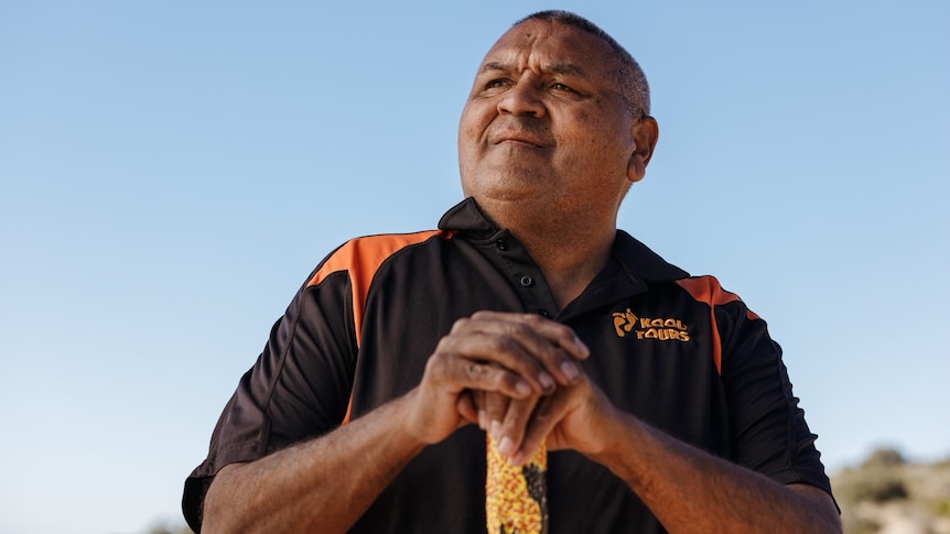 bald, aboriginal man in black polo shirt, hands resting on a staff looking into the distance