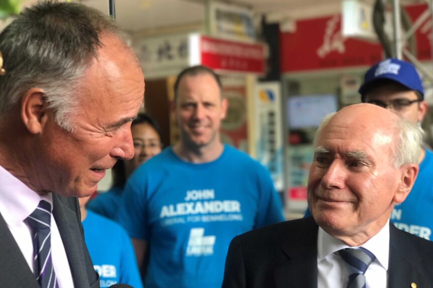 John Howard smiles, tight lipped, looking at John Alexander. Behind them are campaigners wearing blue
