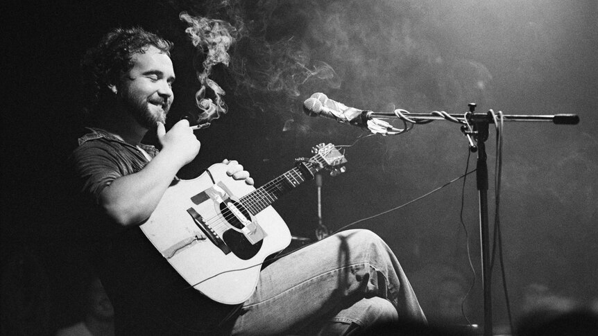 A black and white photo of John Martyn sitting on stage with his guitar. He is smiling and holding a lit cigarette.