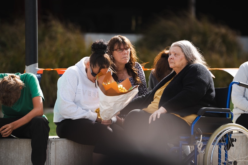 Three women grieve. One is in a wheelchair, another has a bouquet of flowers.