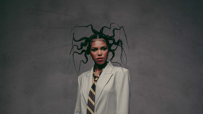 Liyah Knight looks directly into the camera, her hair in wired braids, wearing a grey suit and tie against a grey background