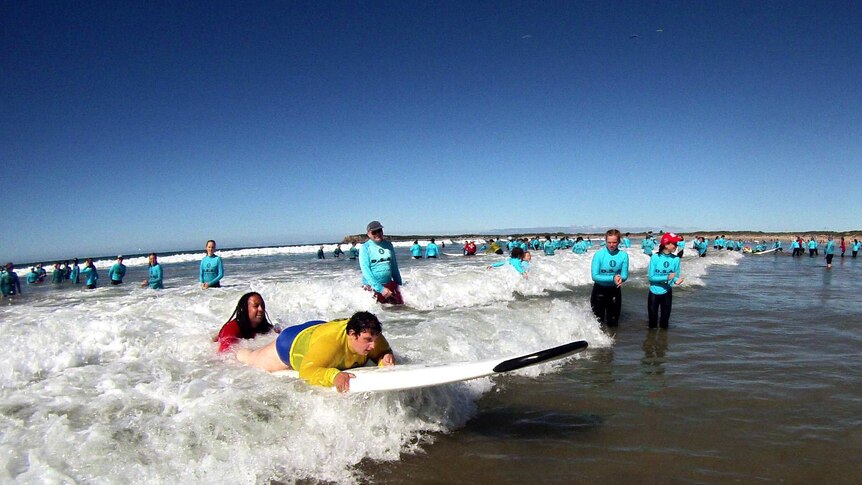 A young woman lying on a surfboard, as it is pushed by a wave, surrounded by clapping people.