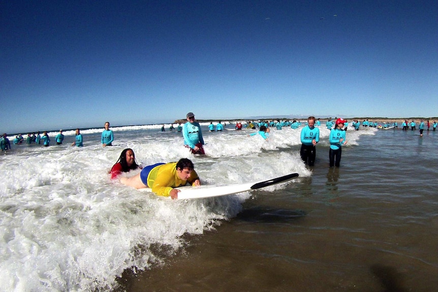A young woman lying on a surfboard, as it is pushed by a wave, surrounded by clapping people.