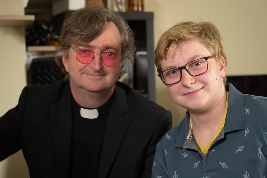 A man wearing a clerical collar with tinted glasses next to his teenage son Elliot who also wears glasses