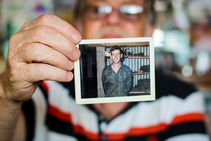 An aged Mick Storen is blurred and the background. He is holding a picture of himself when he was 19.