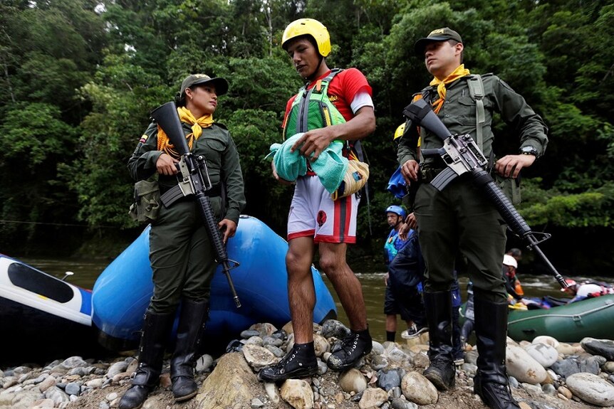 A rafting guide wearing safety gear walks over rocks on a river bank between two armed guards.