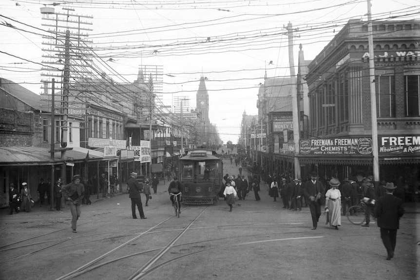 A black-and-white photo of a tram at a bustling city intersection, with streets lined with shops