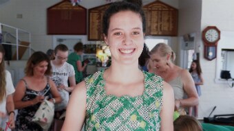 Lily Harrison looks to the camera as she holds to floral bags in her hand. She smiles, and is wearing a green top.