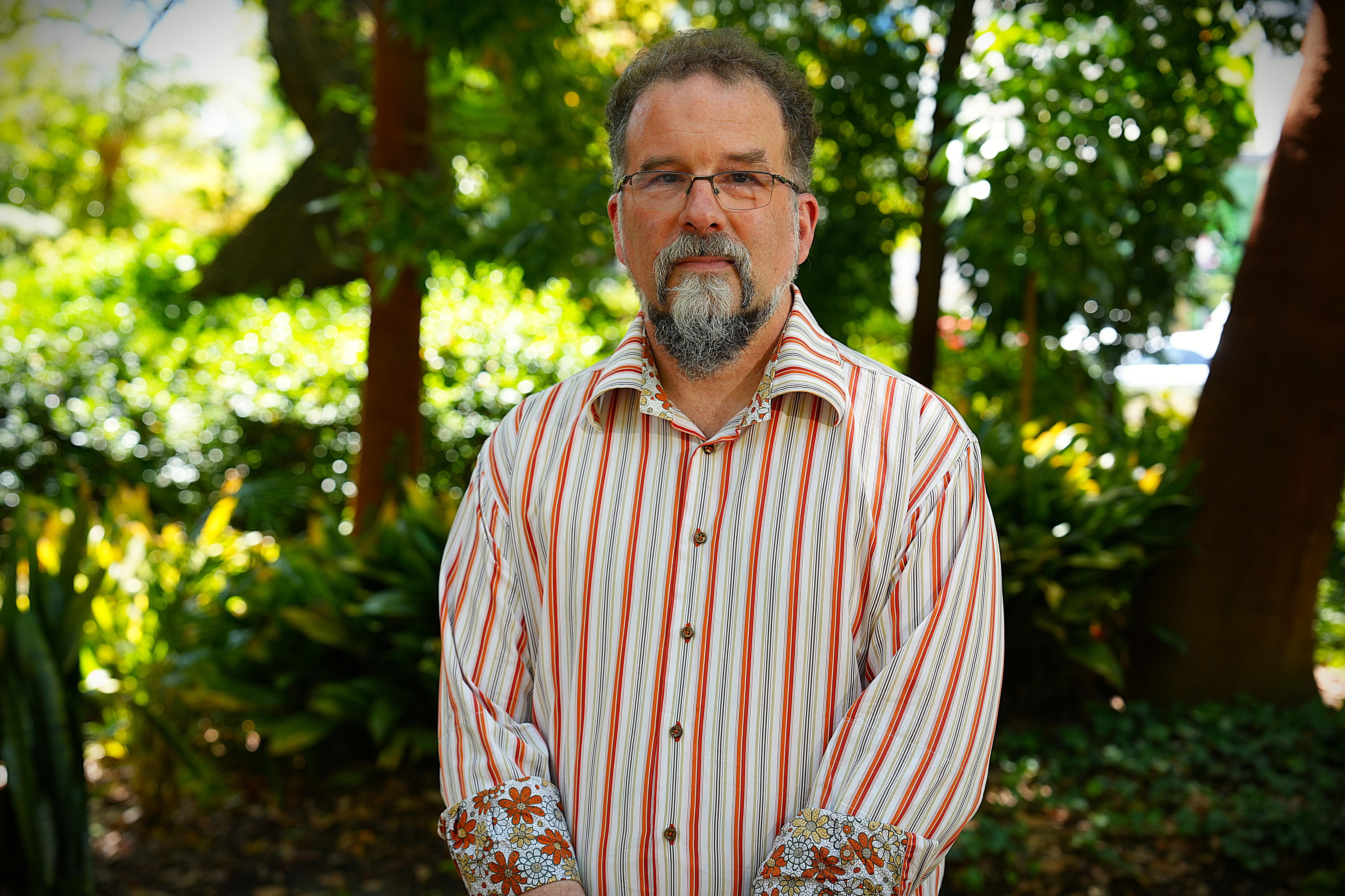 Un hombre barbudo con gafas viste una camisa de cuello de manga larga y se para frente a los árboles. 